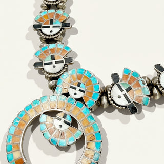 A close-up of a silver-toned Kachepa necklace featuring turquoise, orange, and white inlay details on a light beige background.